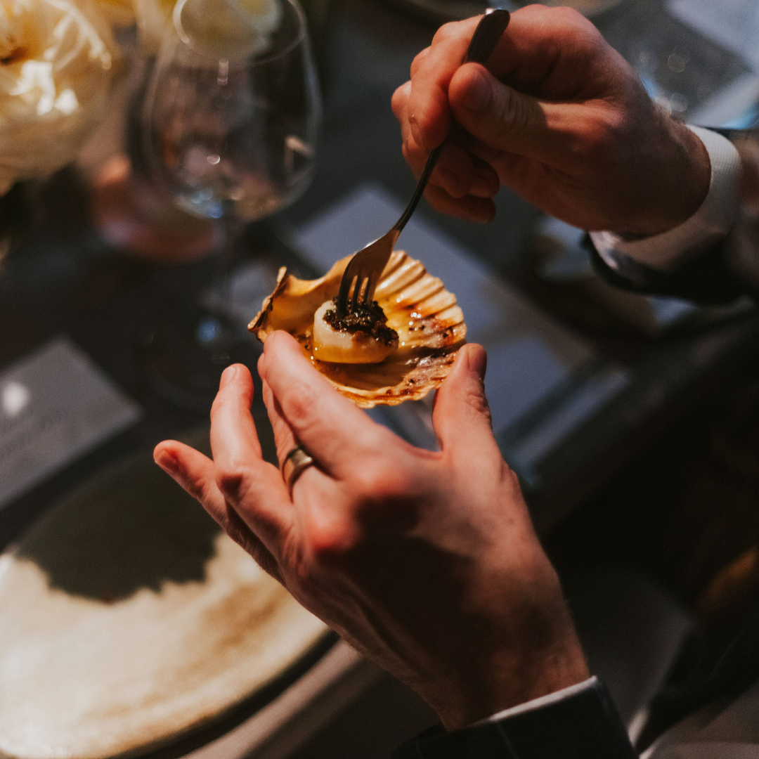 A close-up of a hand holding a scallop shell with a savoury seafood dish, ready for tasting