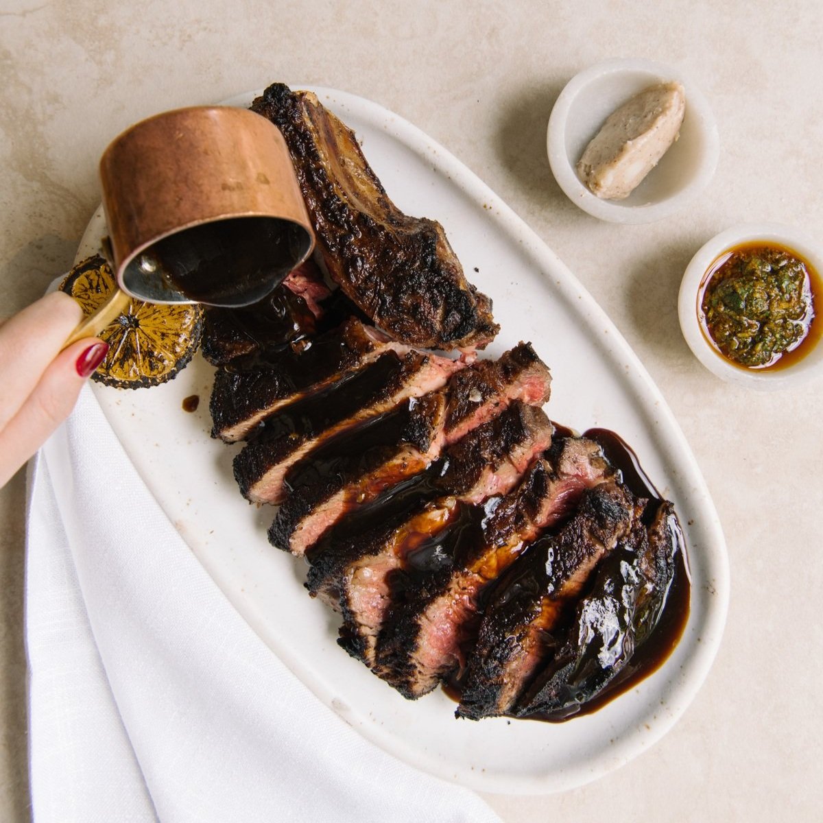 A sumptuous plate of sliced grilled steak, glistening with a rich glaze as it's being poured from a copper jug, accompanied by a charred lemon half, with a small bowl of vibrant green chimichurri sauce and whipped butter on the side