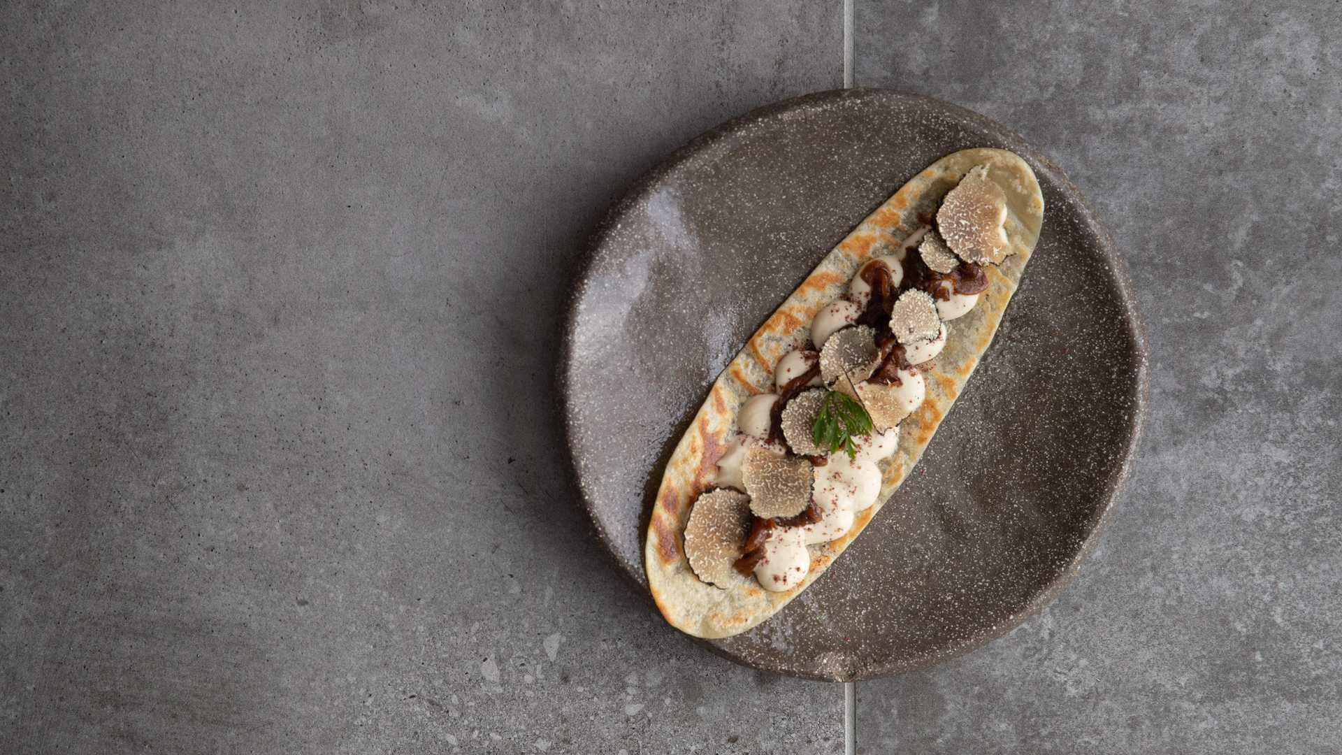 An artistic dish featuring a flatbread elegantly topped with dollops of creamy sauce, thinly sliced truffles, and a light dusting of seasoning, presented on a speckled gray plate against a textured gray background